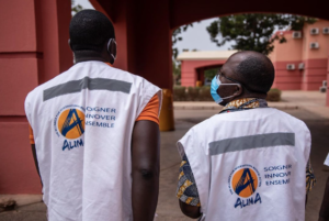 Deux hommes en gilets blancs debout devant un immeuble d’Alima.